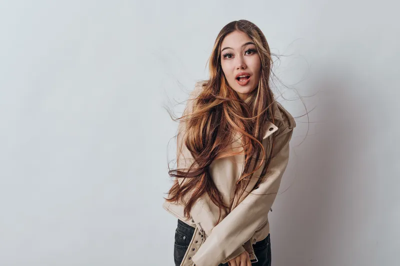 Young woman with long hair and beige jacket posing on white background. Girl with expressive face and flowing hair in casual outfit with black pants and natural light.