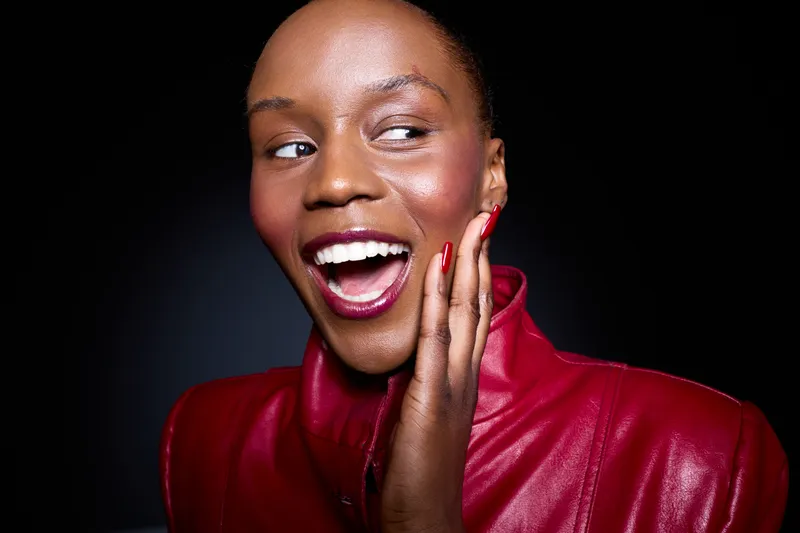 Happy African woman with dark skin laughs joyfully, wearing red leather jacket. She touches her face with manicured red nails against black background. Concept of beauty and happiness.