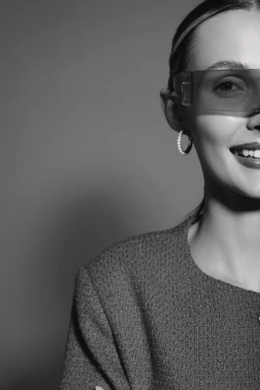 Portrait of a smiling white woman with light skin wearing transparent glasses and textured clothing in black and white.
