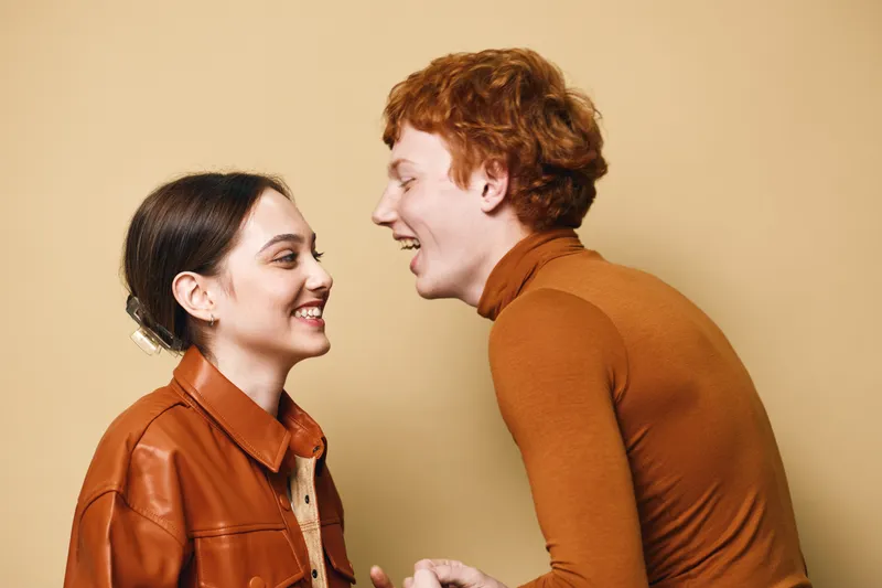 Young man and woman smiling and laughing together indoors, casual clothing, friendly moment captured with beige background in studio environment with happy expressions.