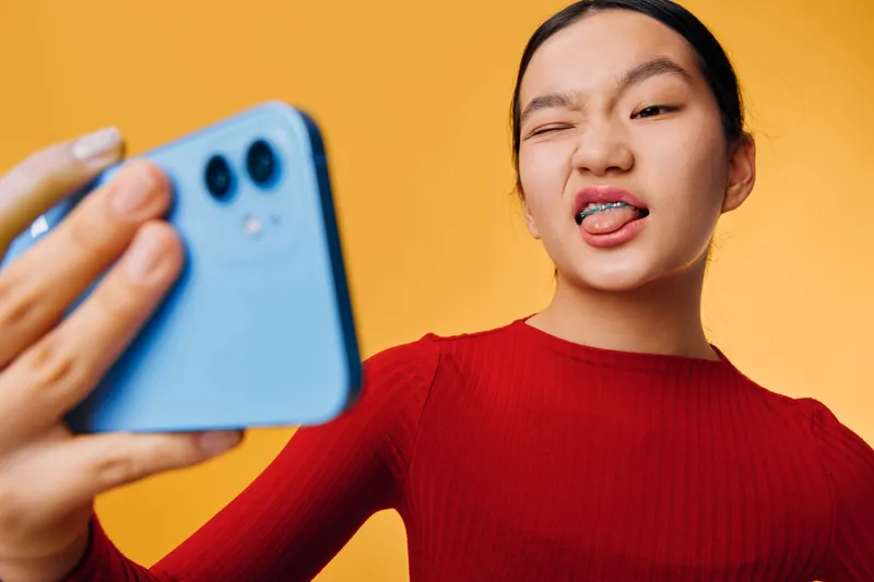 Young woman in red sweater making funny face while holding blue smartphone taking selfie photo against yellow background. Casual portrait of female with playful expression and tongue out.