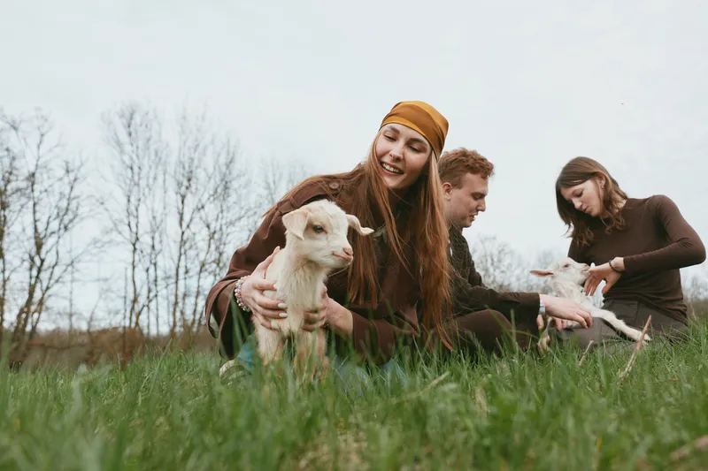 Young woman and couple enjoy a relaxed moment sitting on grass with baby goats in a natural outdoor setting during a cool day in casual autumn clothes.