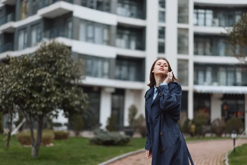 Young woman in blue trench coat talks on smartphone outdoors in city park near modern apartment building, casual style, autumn season, thoughtful expression.