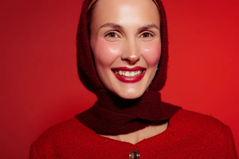 Smiling woman in red sweater and matching headscarf poses against red background. Her glowing skin and bright lipstick create a vibrant and joyful mood in this close-up portrait.