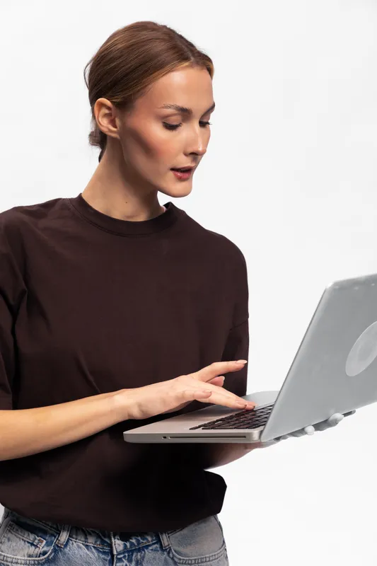 Young woman with red hair wearing dark sweater stands holding open laptop looking at screen with focused expression on white background. Concept of technology, work, and concentration.