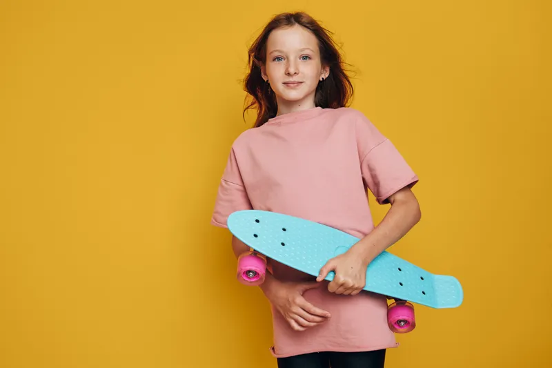 Girl in a pink t-shirt holding a blue skateboard with pink wheels stands against a yellow background. Casual youth concept with vibrant colors and confident expression.