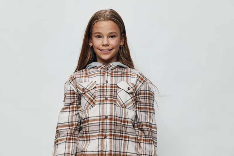 Child girl with long hair wearing plaid jacket standing against plain background smiling at camera with confident and happy expression in studio portrait.