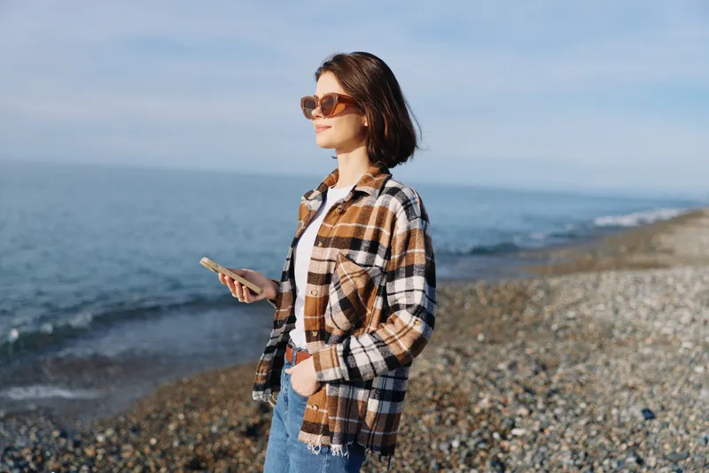 Woman on a pebble beach with phone, wearing sunglasses and a plaid shirt, enjoying coastal light in a casual outdoor portrait for travel, lifestyle and nature themes.