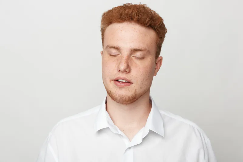 Young redhead man with closed eyes and light skin wearing white shirt stands against plain background. Concept of relaxation, calmness, meditation, and peaceful moment in studio portrait.