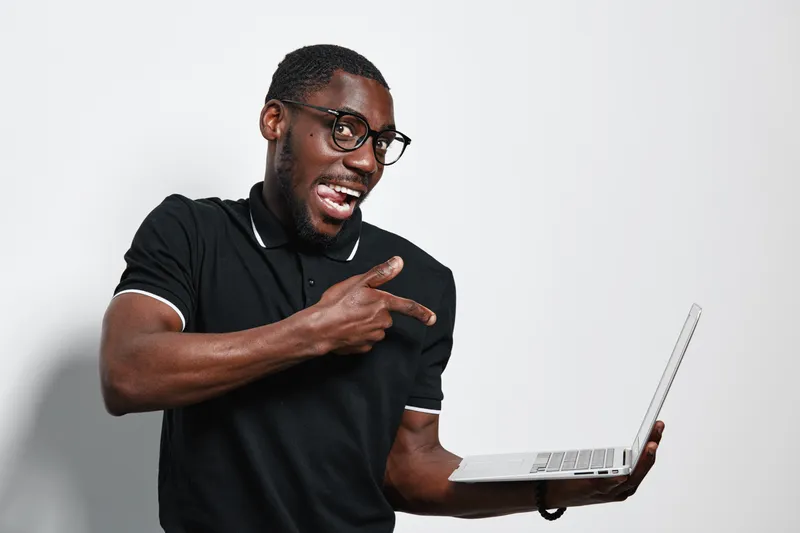 African American man with dark skin and glasses holding laptop and pointing finger with excitement on white background. Casual black shirt, technology, communication and digital device.