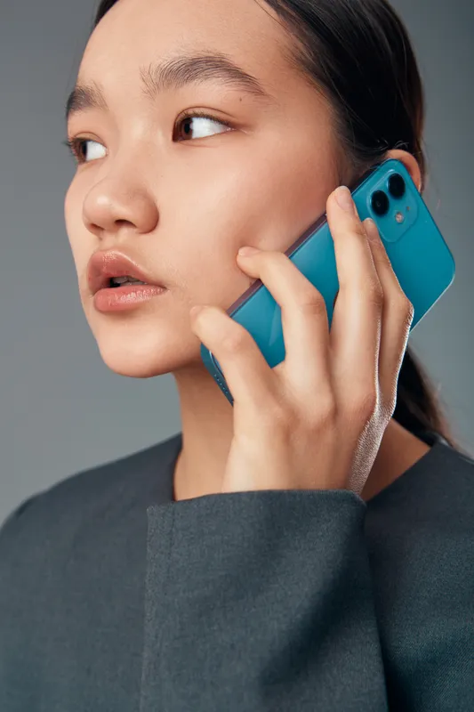 Woman holding blue smartphone near ear in closeup portrait, looking sideways with serious expression in casual outfit on gray background, communication and technology concept.