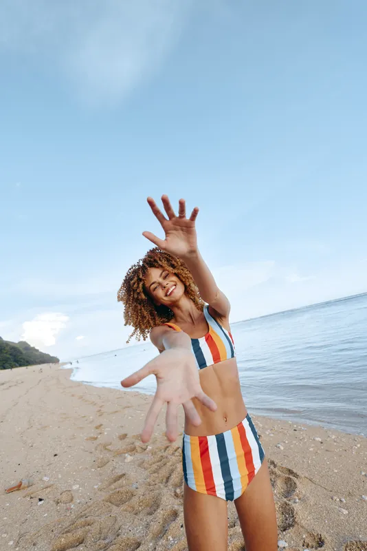 Smiling woman in a striped bikini stands on a sunny beach reaching toward the camera, creating a vibrant, carefree mood with bright colors, warm light, and outdoor summer freedom.