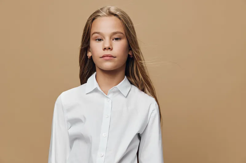Girl with long hair in white shirt standing with neutral expression against beige background for portrait, youth, casual style and confident mood indoors.