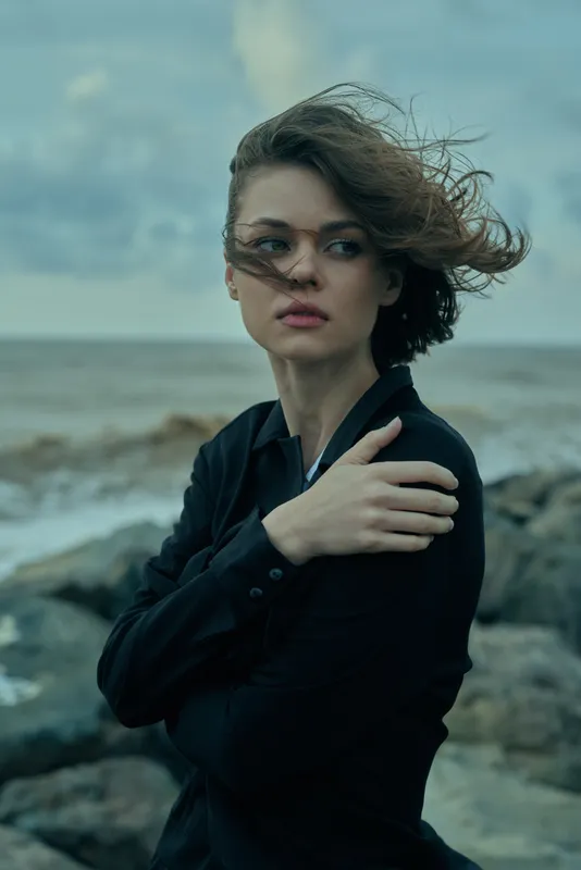 Dramatic, windswept coastal scene with a woman in a black top, flowing hair, strong gaze, and folded arms, rocks and ocean under a stormy sky, moody fashion mood, cinematic lighting and atmospheric