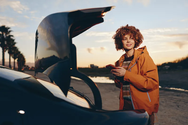 Smiling woman lifestyle stands by an open car door at sunset on a quiet road, holding a phone and facing the camera. Casual jacket and warm evening light create a travel mood.