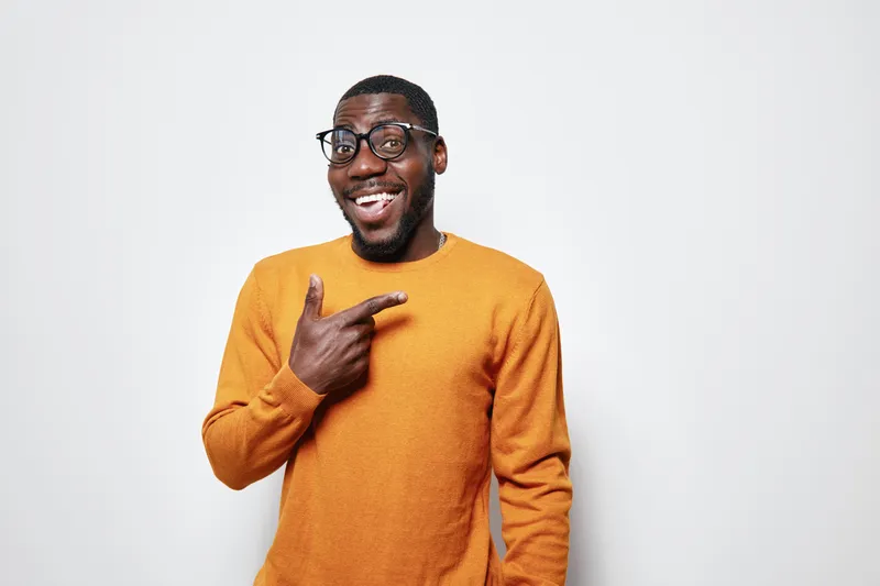 African American man with dark skin wearing glasses and orange sweater smiling and pointing finger to side on white background. Positive expression and casual style portrait.