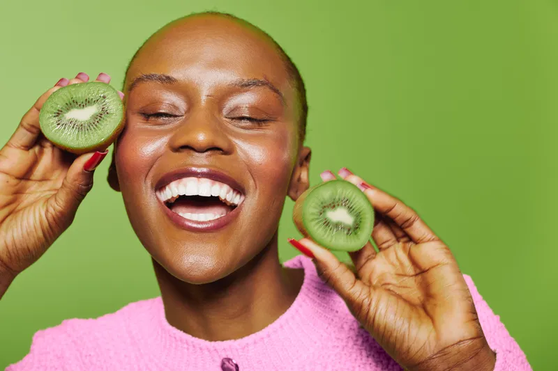 Happy smiling African woman with dark skin holds two halves of fresh kiwi fruit close to face on green background. Concept of healthy lifestyle, natural food, and joyful mood.