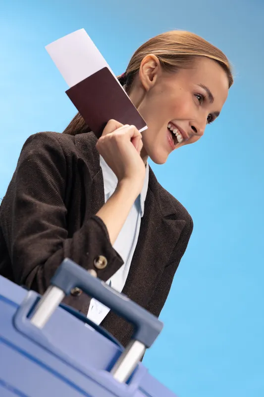 Happy woman traveler holds passport and ticket near blue suitcase. She smiles and looks away with excitement. Concept of travel, vacation, tourism, and journey planning.