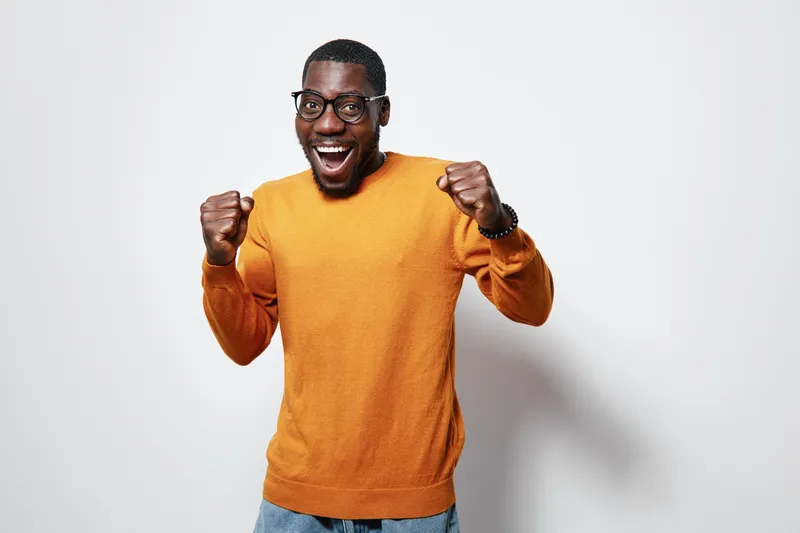 Excited African American man with dark skin wearing glasses and orange sweater celebrating success with clenched fists and joyful expression on white background.