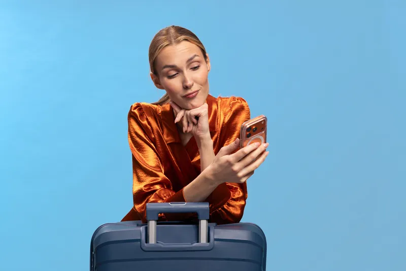 Young woman in shiny orange blouse leans on blue suitcase while looking at smartphone with thoughtful expression. Concept for travel planning, communication, and modern lifestyle.