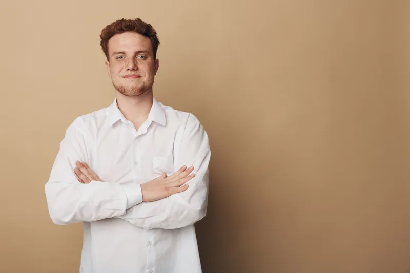 Confident young man with curly hair wearing white shirt stands with crossed arms against beige background. Portrait of casual male model with relaxed and self-assured expression.