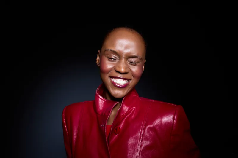 Close-up of happy African woman with dark skin wearing red leather jacket smiling with closed eyes against black background. Concept of joy, fashion, confidence, and beauty.