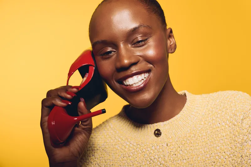 Smiling dark skinned African woman holds red high heel shoe near face, wearing beige sweater on yellow background. Concept for fashion, beauty, happiness, and style portrait.