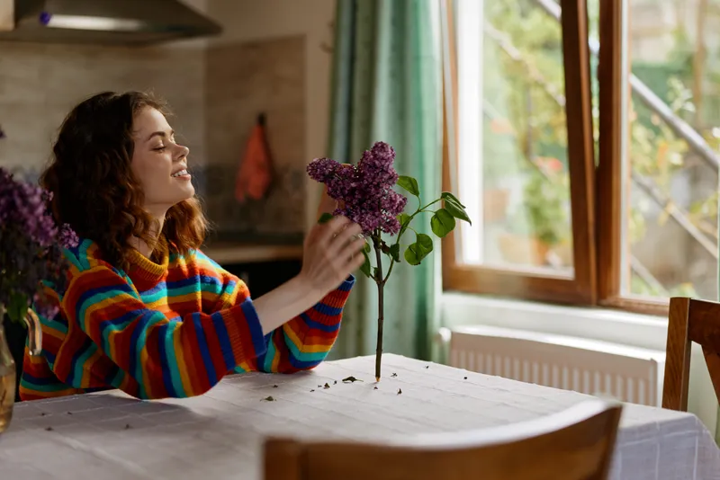 A woman in a colorful striped sweater tends to a purple flower at a dining table, bright window light fills a cozy home space, mood of calm and domestic care.