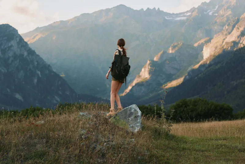 Young woman with a backpack stands on a rock in a mountain field enjoying the scenic view during a hiking adventure at sunset.