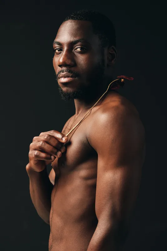 African man with dark skin and muscular body posing shirtless in creative conceptual style studio portrait with dramatic lighting and confident expression on black background.