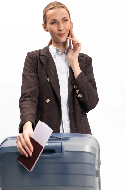 Young woman in brown blazer holding passport and phone while pulling large suitcase. Business travel concept with confident female traveler ready for trip. Isolated on white background.