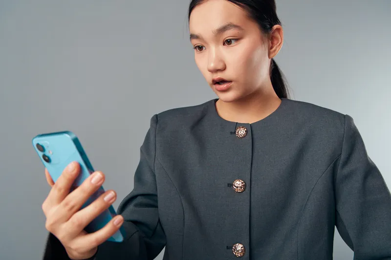 Young woman in gray jacket looks surprised holding smartphone with open mouth and focused eyes on gray background. Female person reacting with mobile device and unexpected news.