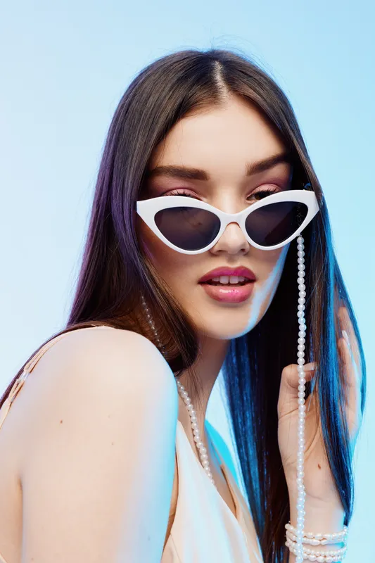 Young woman with long hair wearing white sunglasses and pearl accessories poses confidently against a light background. Concept of beauty, fashion, and style.