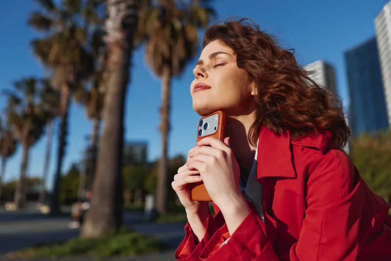 Young woman outdoors enjoying sunlight with eyes closed holding smartphone in red coat, relaxed expression, palm trees and urban background.