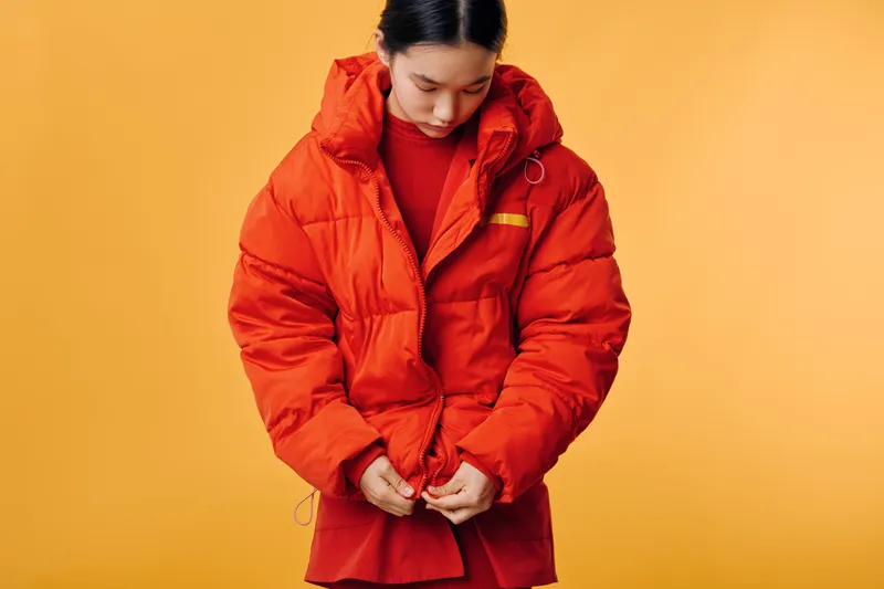 Woman wearing red puffer jacket and red sweater standing against yellow background. Young adult with dark hair looking down while adjusting coat zipper in studio setting.