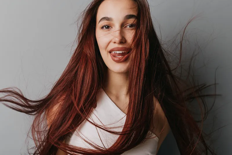 Woman with long red hair smiling and sticking out tongue in studio. Female model with flowing hair wearing white top on gray background with playful expression and natural makeup.
