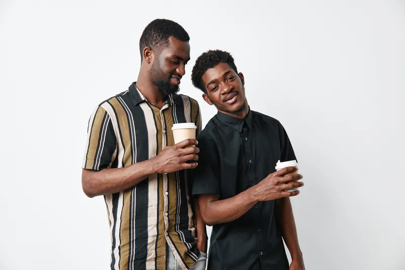 Two young black men with dark skin holding coffee cups and smiling together in casual clothing on white background. Friendship, relaxation and positive mood indoors.