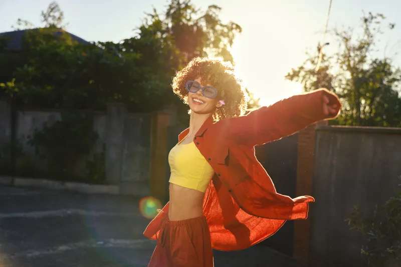 Joyful woman with curly hair dances outdoors in a bright yellow top and red jacket, embodying a carefree lifestyle in a sunlit retro mood