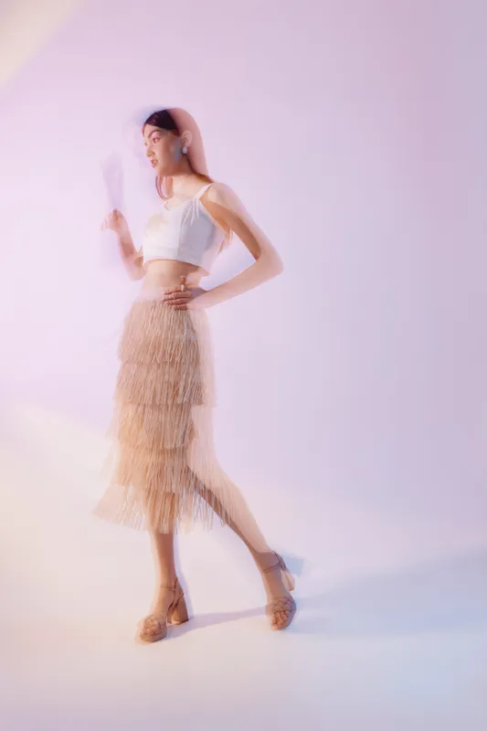 Young woman in fashionable outfit poses with hand on hip and holding a fan. Soft lighting highlights beauty and style in a minimal studio setting. Concept of woman beauty fashion.