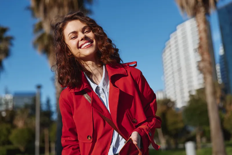 Smiling woman with curly hair wearing a red coat outdoors in a sunny urban park, bright sky and trees in the background, casual lifestyle and fashion vibe.