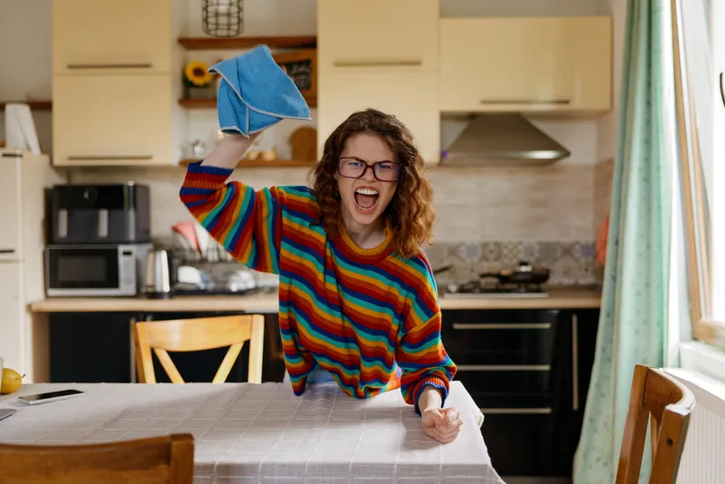 Young woman with curly hair cleans a bright kitchen, wiping the table with a blue cloth and laughing, a candid home moment filled with sunlight and everyday comfort.