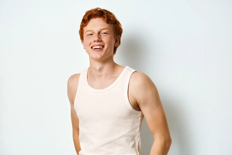 Young man with red hair and freckles wearing white tank top smiling confidently on white background in casual style portrait.
