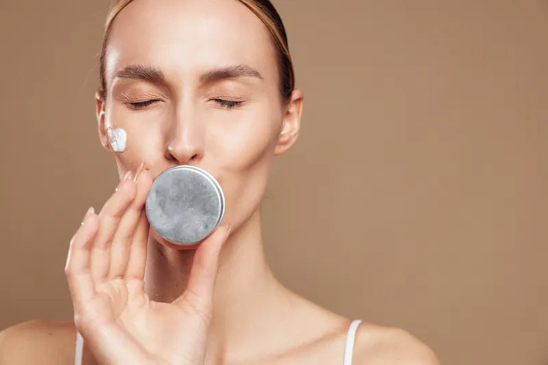 Woman with closed eyes applying face cream on cheek holding a round cosmetic container close to lips. Concept of skincare, beauty, wellness, and self-care on neutral background.