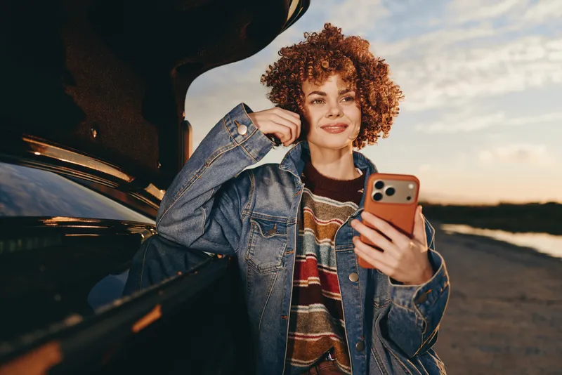 Woman lifestyle smartphone denim car sunset portrait curly hair, smiling, holding phone near open car door, casual streetwear, warm golden light, outdoors evening concept