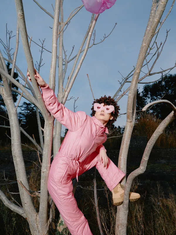 Person in a pink jumpsuit climbs a bare tree outdoors, a pink balloon drifting above, creating a whimsical and playful scene ideal for creative, lifestyle or fashion stock with natural light and