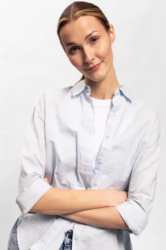 Confident young woman in casual white shirt stands with arms crossed against white background. Portrait of natural beauty, relaxed mood, and casual style for lifestyle and fashion concept.