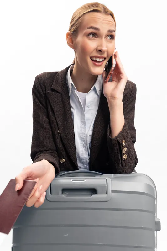 Smiling woman talking on smartphone holds passport with suitcase in foreground. Business casual dressed traveler ready for trip isolated on white background. Concept of travel and communication.