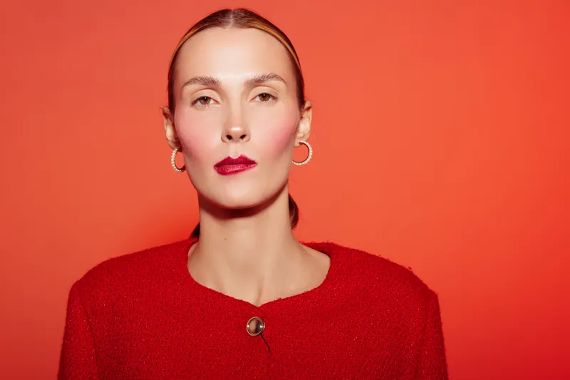 Portrait of confident woman with slicked back hair wearing red sweater and hoop earrings against orange background. Concept of fashion, beauty, elegance, and minimalistic style.