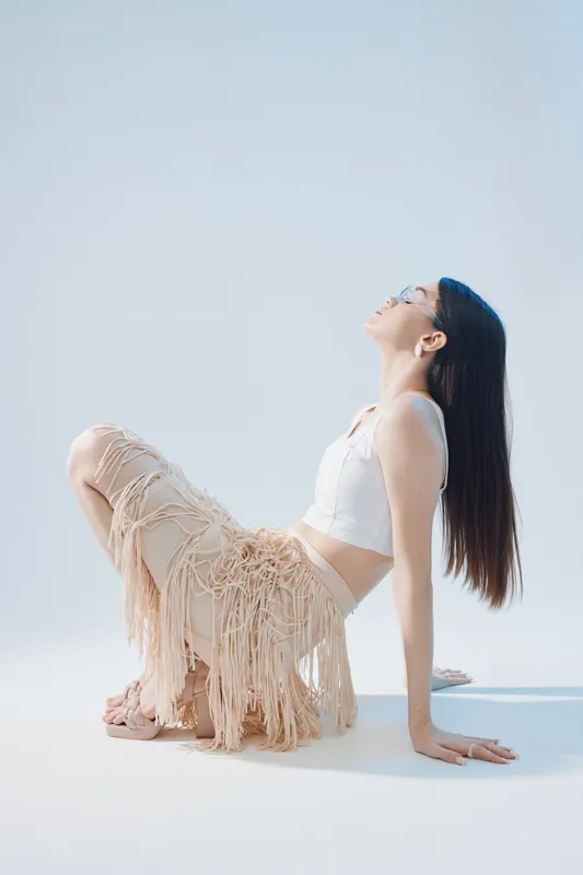 Young woman in a white top and fringed skirt poses sitting on the floor with her head tilted back. Bright studio lighting highlights her beauty and fashion style. Concept for woman beauty fashion.