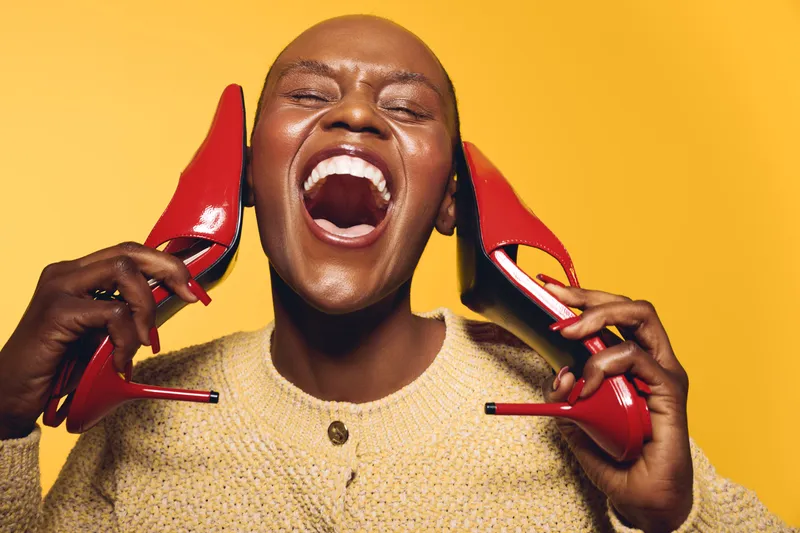 Happy dark skinned African woman with shaved head holds red high heel shoes near face, laughing with closed eyes on yellow background. Concept of joy, fashion, and beauty with colorful style.